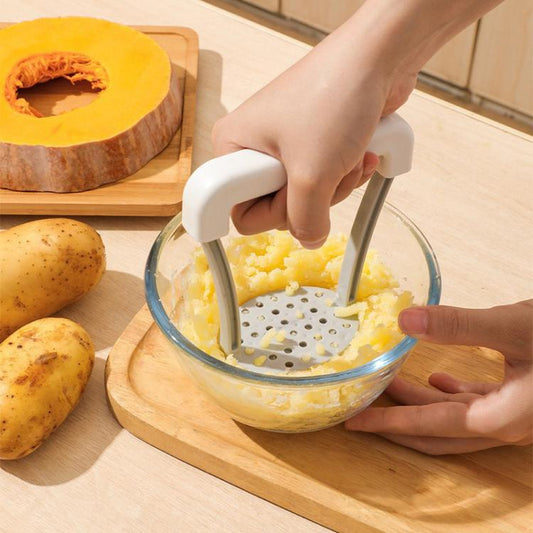 Manual potato masher pressing yellow potatoes in a glass bowl; white handle and plate