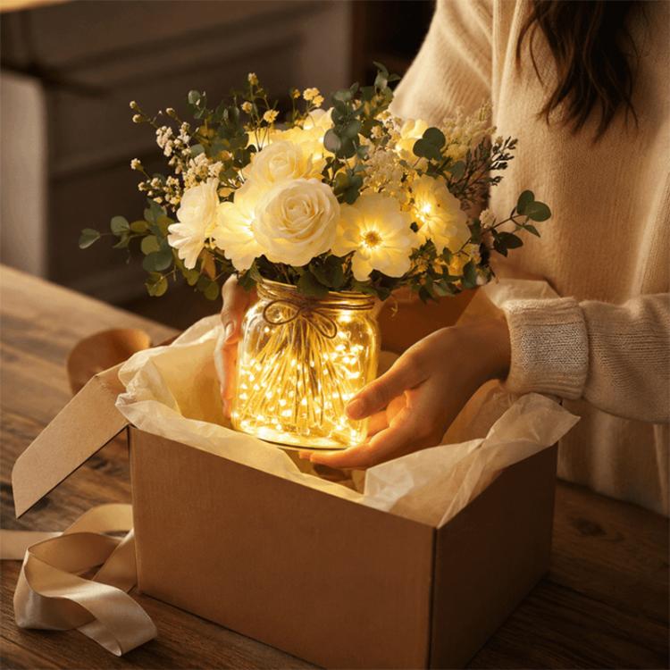 LED faux flower bouquet in glass bottle being placed in gift box, warm lights and white blooms