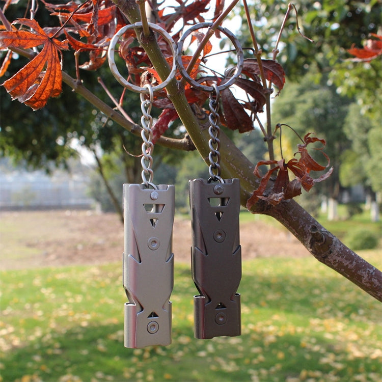 Two double-tube survival whistle keychains hanging outdoors, light and dark metal bodies