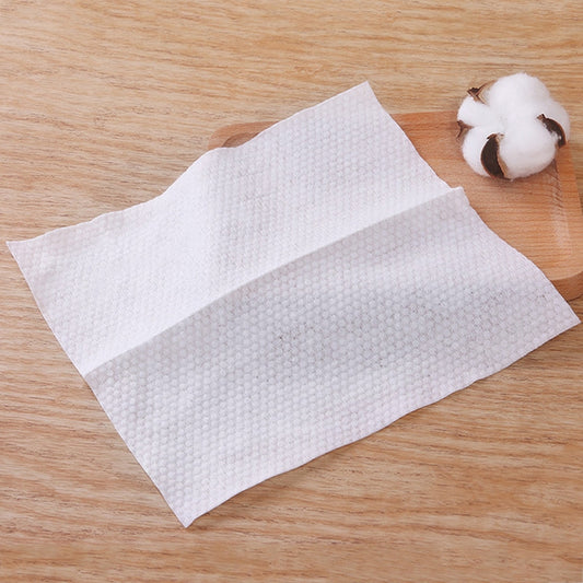 Two white textured disposable cotton face towels on wooden surface with small cotton ball