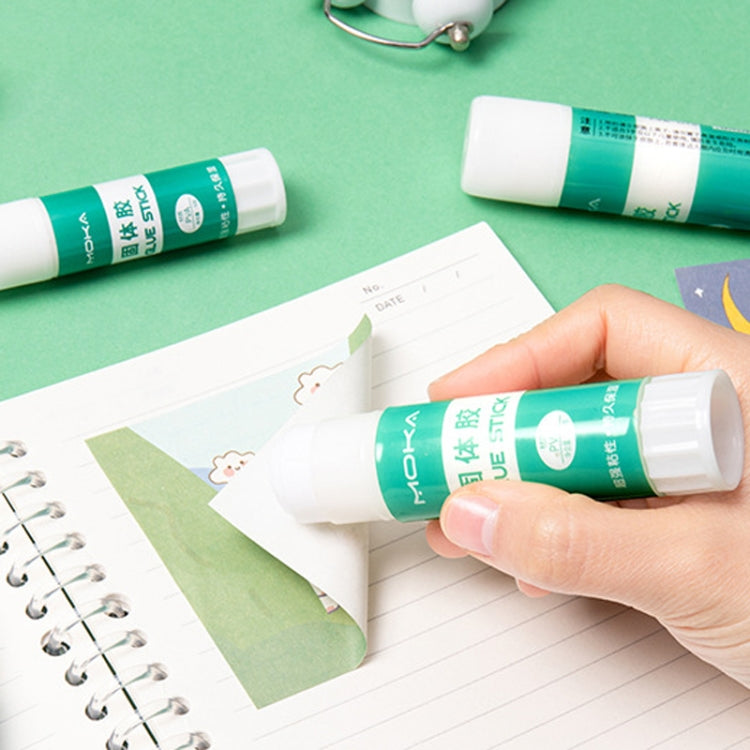 Solid glue stick applying glue near notebook pages; green label and white cap visible.