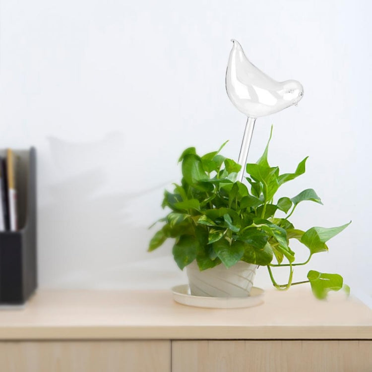Glass bird watering stake inserted into green potted plants, clear spout above