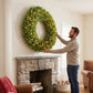 Green pre-lit Christmas wreath with warm LED lights being mounted on a wall above a fireplace.