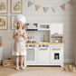 Child in chef hat and apron standing by white kids play kitchen with gold faucet