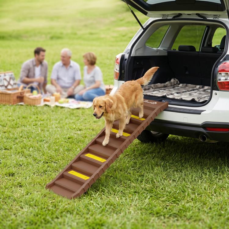 Brown foldable dog ramp with yellow step strips, dog walking from open car trunk to grass