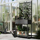 Black outdoor wicker planter with trellis, green plants and lower shelf in a greenhouse setting
