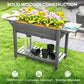 Gray wooden raised planter box with flowers, watering can, and bottom shelf on wheels