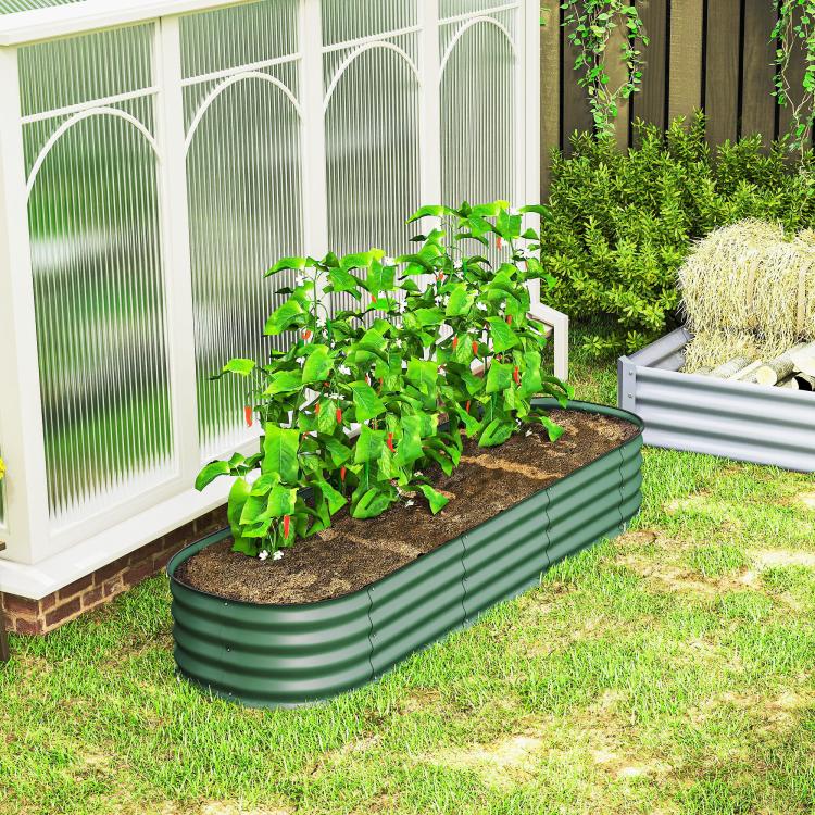 Green metal raised garden bed planter with leafy plants beside a greenhouse structure
