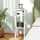White 2-tier side table with drawer and open shelf next to a sofa and plant pot.