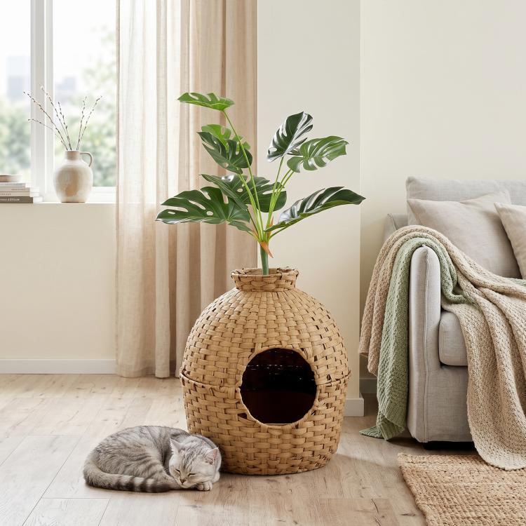 Rattan-style cat litter box with faux plant, round doorway, and gray cat resting nearby