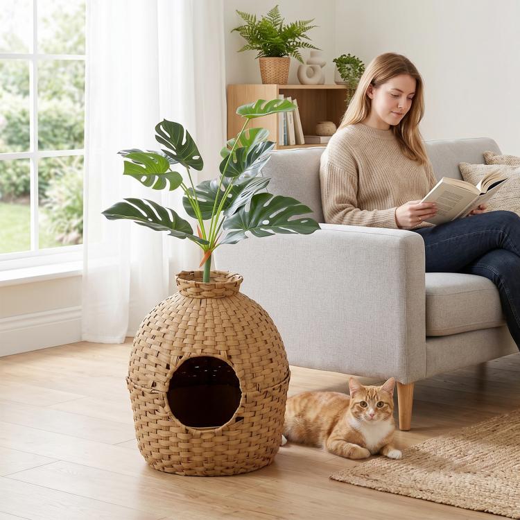 Rattan-style cat litter box with faux plant top and round front doorway next to orange cat
