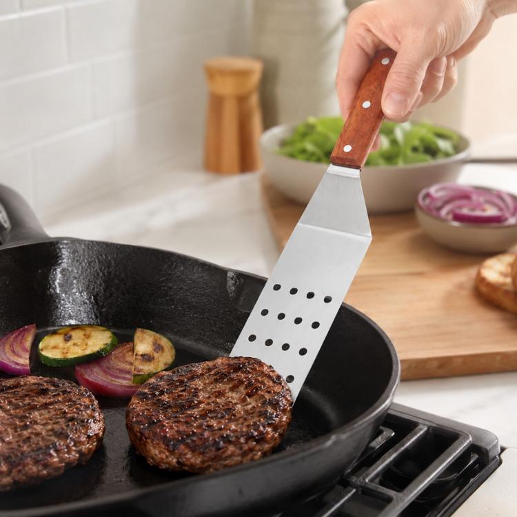 Stainless steel slotted spatula with wooden handle, lifting grilled meat from a skillet