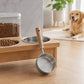 Silver stainless steel pet food measuring scoop with wooden handle on a wooden tray