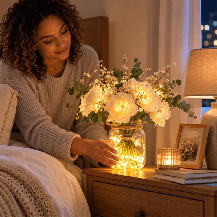 LED faux flower bouquet in clear glass bottle on bedside table, white flowers glowing warm