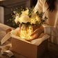 LED faux flower bouquet in glass bottle being placed in gift box, warm lights and white blooms