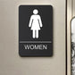 Black Braille sign with raised female icon, tactile dots, and WOMEN label.