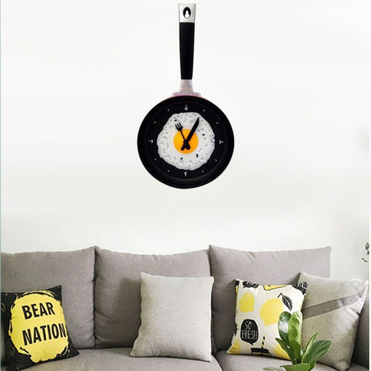 Frying pan-shaped wall clock with black rim, orange yolk center and fork-and-spoon hands