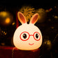 Smiling bunny LED lamp glowing warm light, red glasses, rounded silicone body