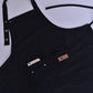Black kitchen apron close-up showing logo patch, buttons, and pockets.