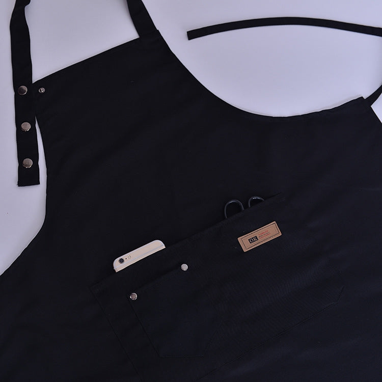 Black kitchen apron close-up showing logo patch, buttons, and pockets.