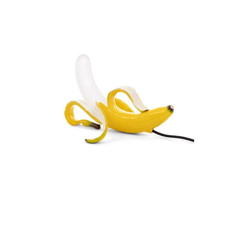 Banana table lamp with yellow banana body and white bulb, black cord lying across