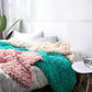 Chunky wool knit throw blankets in teal, blush pink, and cream stacked on a bed
