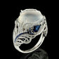Silver-tone statement ring with oval white cabochon, dark blue gem, and feather band