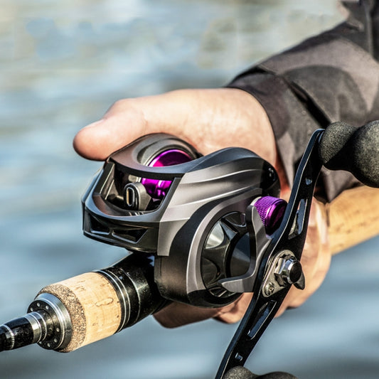 Hand holding baitcasting fishing reel with purple spool and black handle grips.