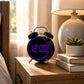 Black twin-bell digital alarm clock with purple glowing ring on a bedside table