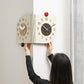Dual-dial corner wall clock held by hands; beige and dark brown faces with red knob