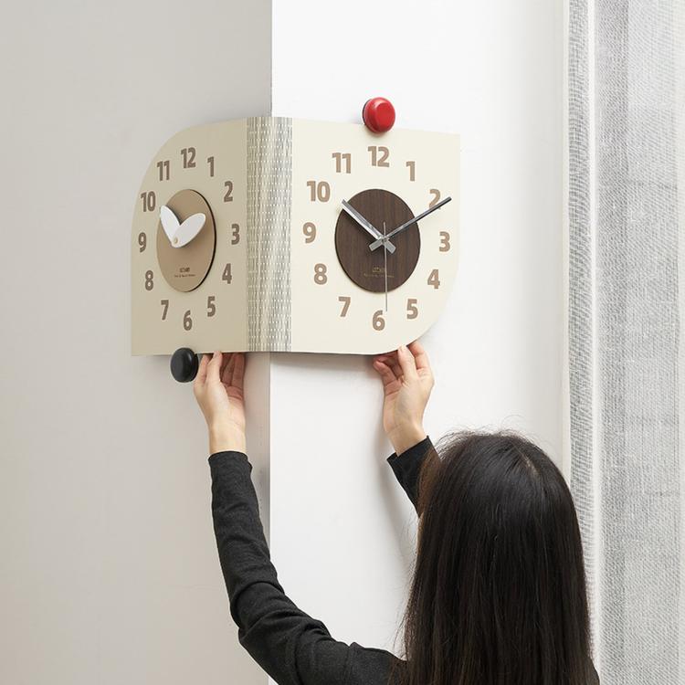 Dual-dial corner wall clock held by hands; beige and dark brown faces with red knob
