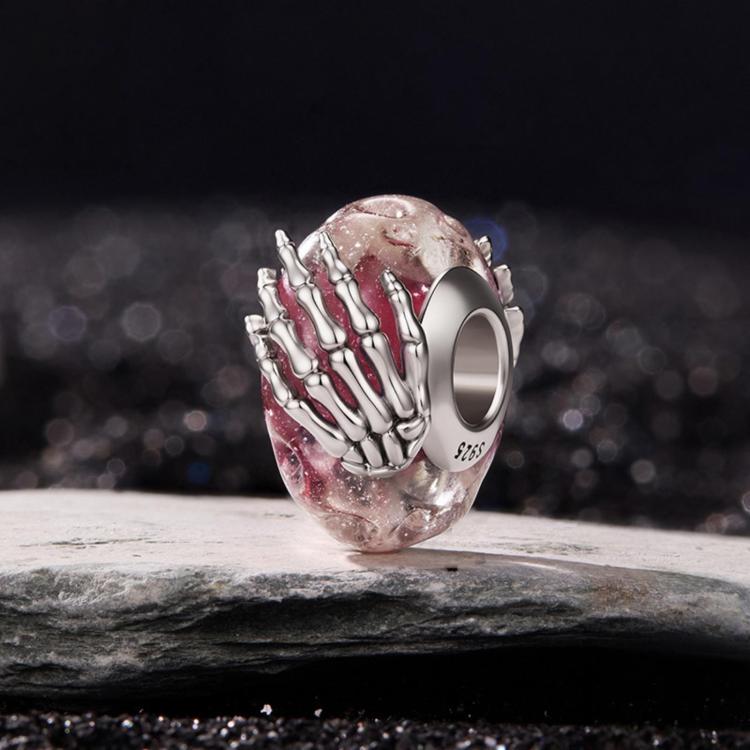 Pink glitter glass bead charm with silver skeleton hand; side view on gray surface