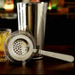 Stainless steel ice trap strainer with perforated disc and long handle, shown on bar setup